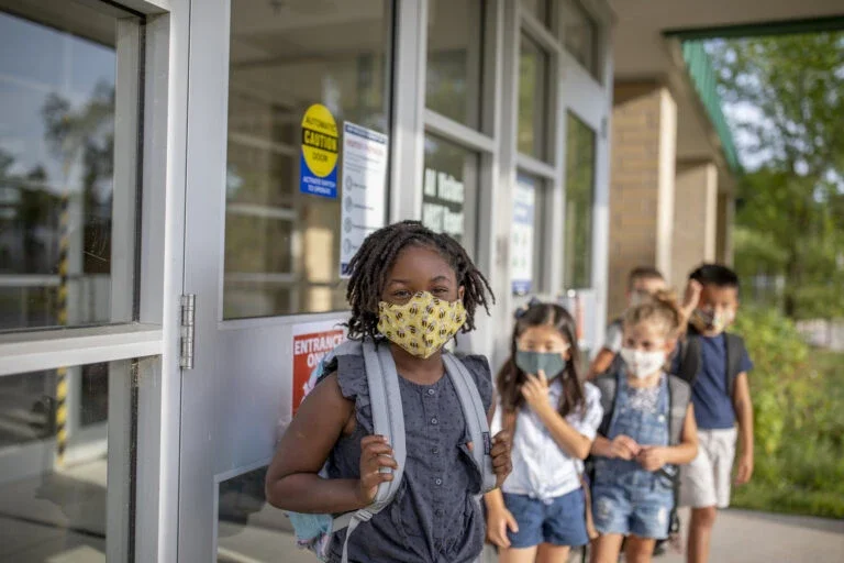 young boys and girls lined up outside of school wearing backpacks and masks.